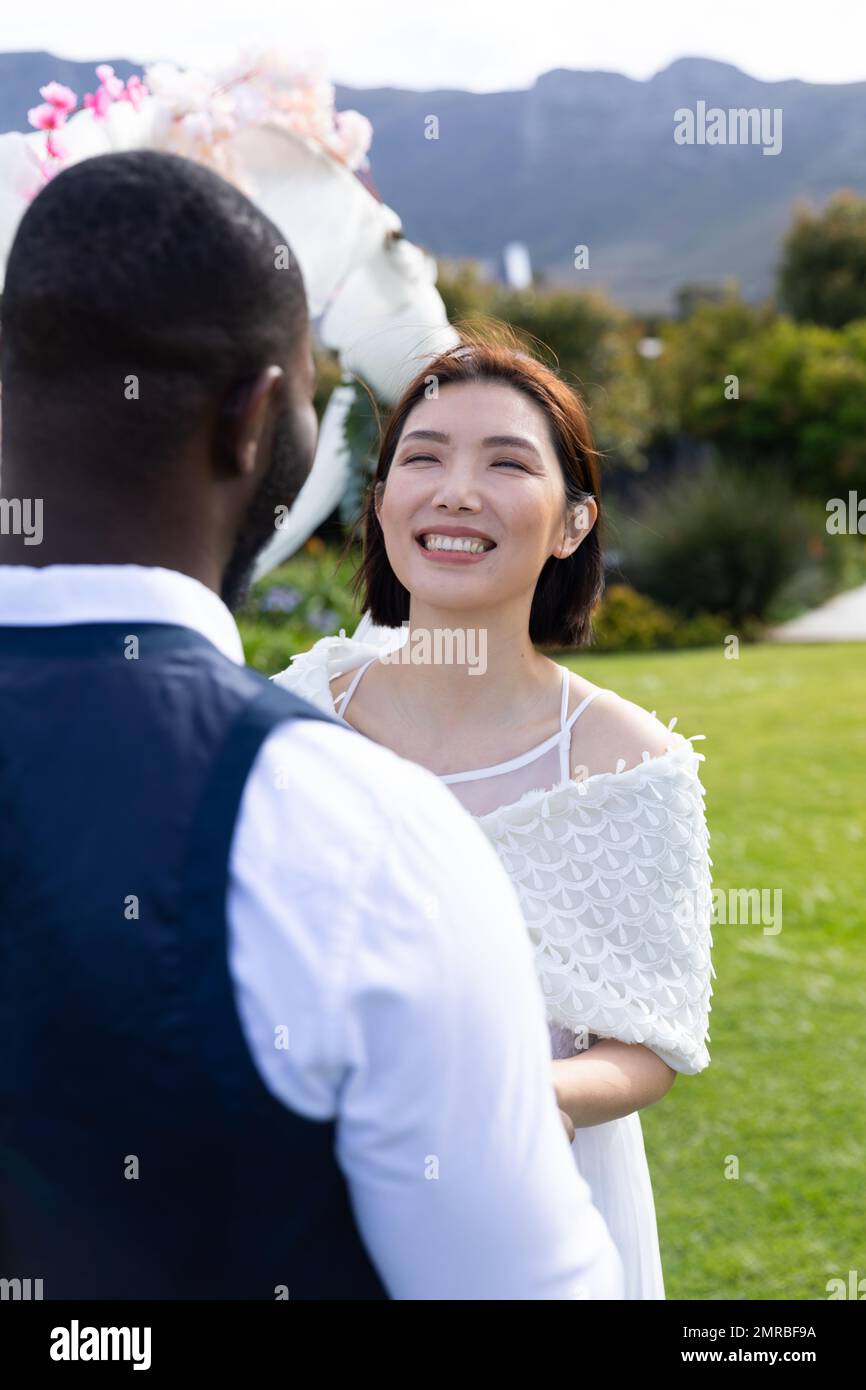 Verticale di felice sposa asiatica sorridente a sposo diverso a nozze all'aperto, copia spazio Foto Stock