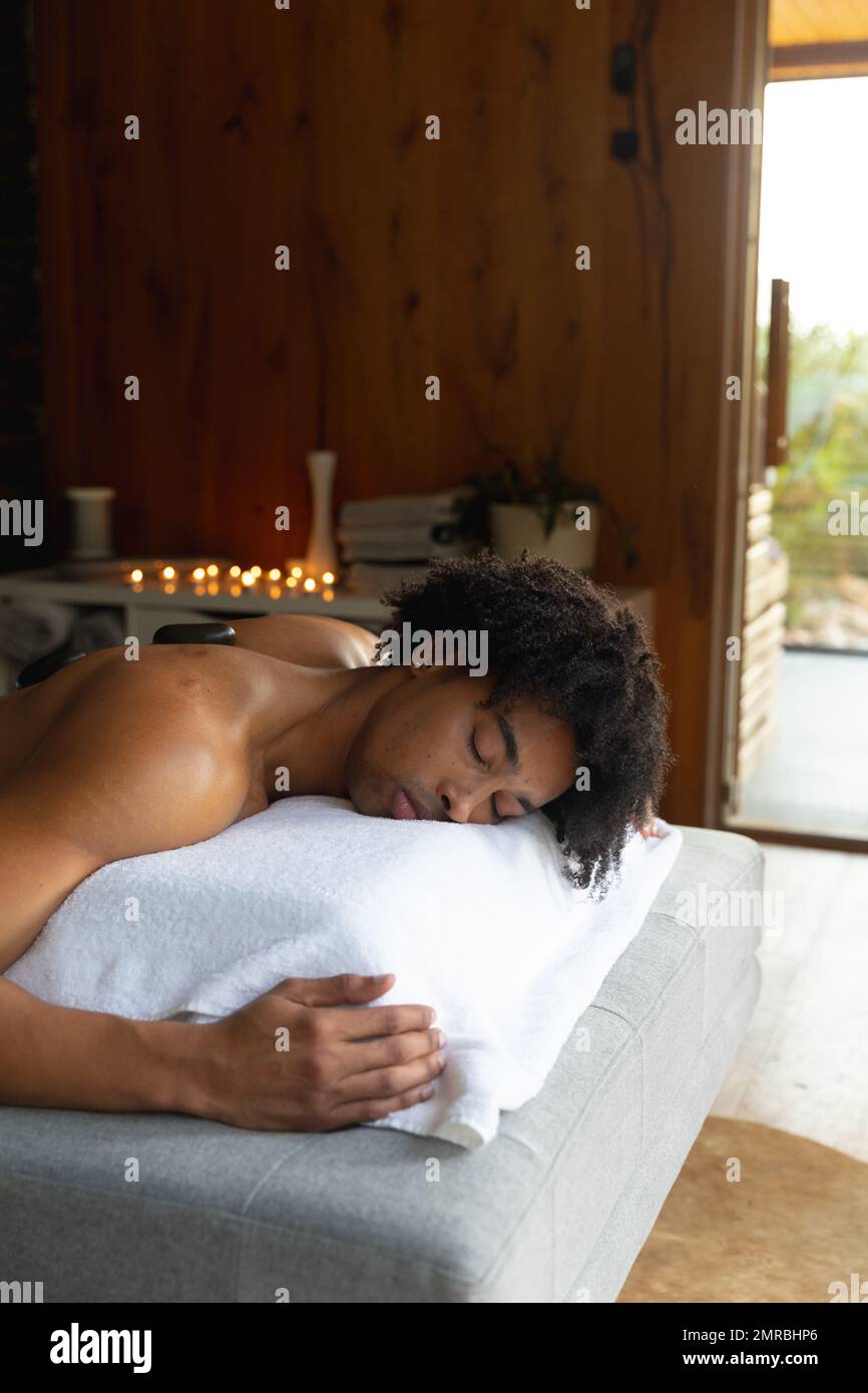 Verticale di uomo biraciale rilassato sul tavolo da massaggio con trattamento pietre calde presso la spa, spazio copia Foto Stock