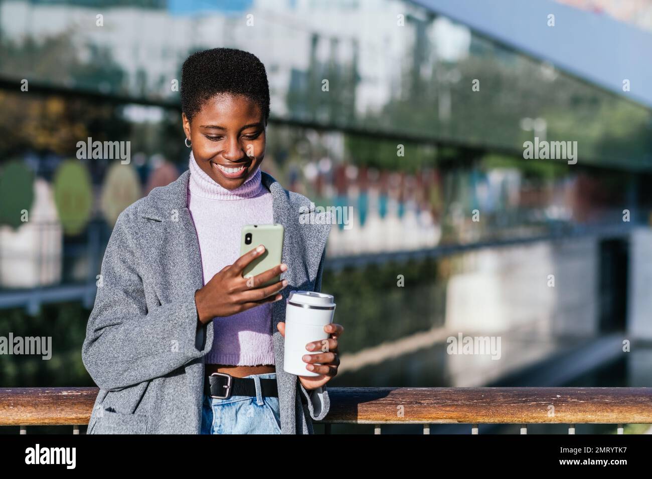 Ragazza afro-americana in elegante outerwear con tazza di caffè senza sprechi sorridendo e navigando sui social media sul cellulare. È in piedi sul ponte pedonale sullo sfondo sfocato della strada cittadina Foto Stock