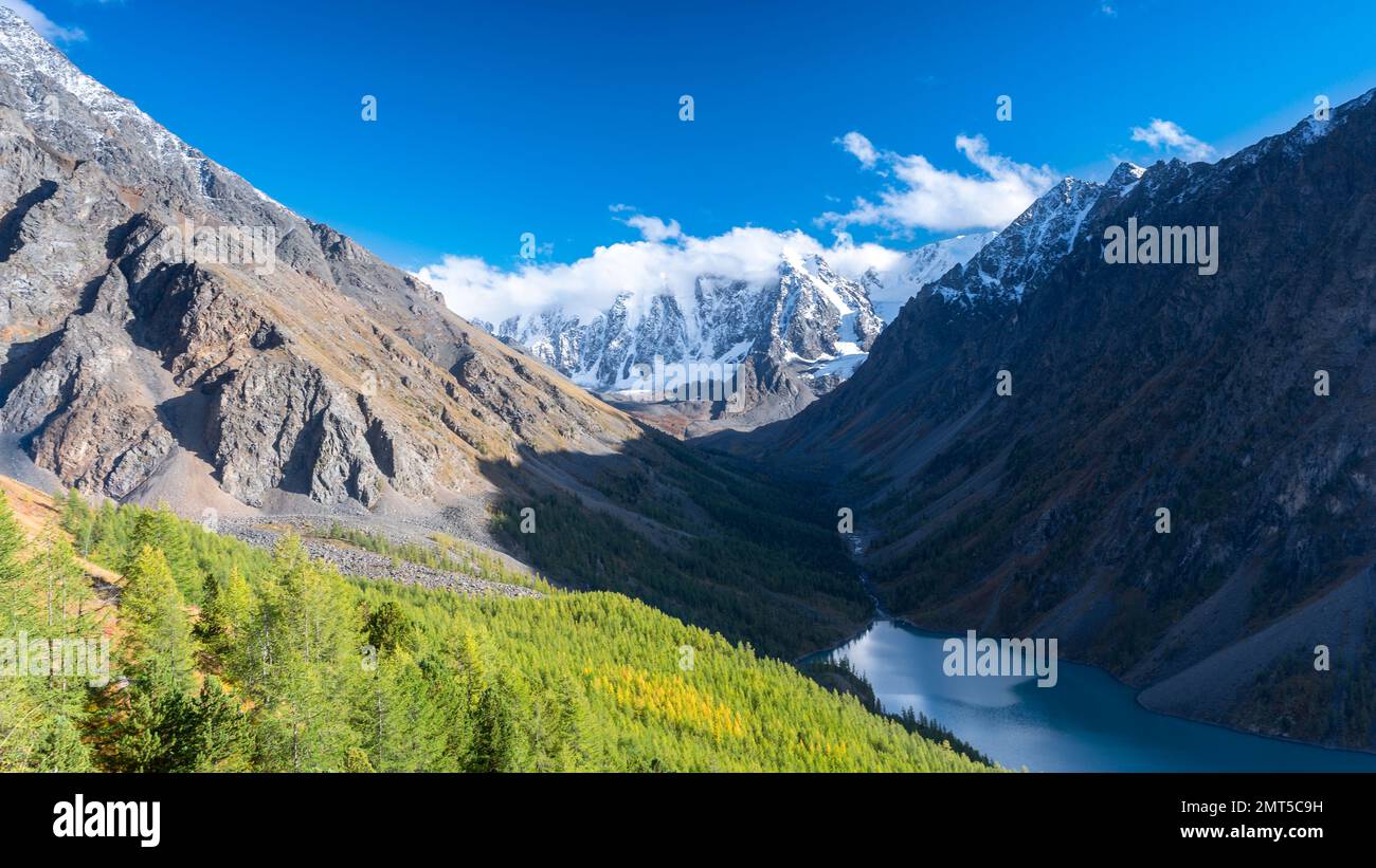 Lago di montagna Shavlinskoye all'ombra senza sole tra le rocce di pietra dietro la foresta sullo sfondo di cime innevate con ghiacciai Foto Stock