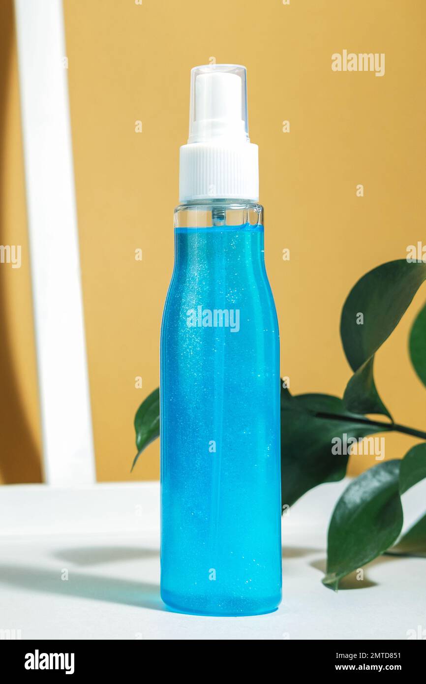 Nebbia di capelli. Mockup di un contenitore cosmetico con un flacone spray, con un liquido blu. Su sfondo bianco arancione con ombre e foglie dure. Vertica Foto Stock
