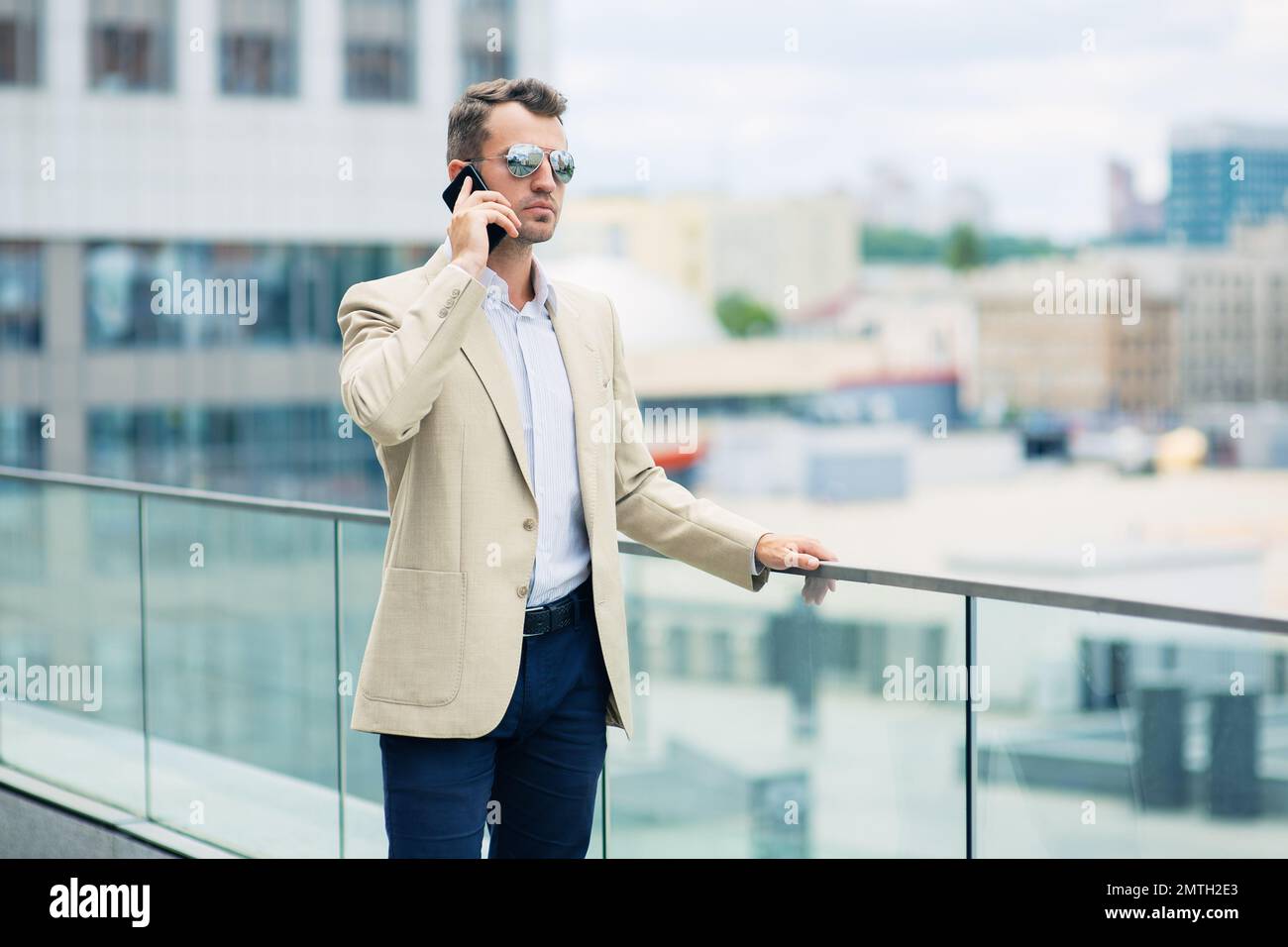 Elegante sicuro giovane e impegnato uomo d'affari in una giacca elegante e occhiali da sole che parlano sul telefono cellulare mentre si è in piedi all'aperto nel centro città Foto Stock