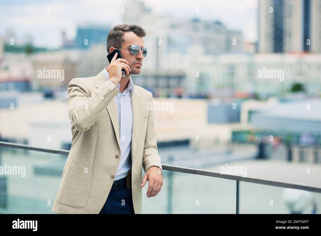 Elegante sicuro giovane e impegnato uomo d'affari in una giacca elegante e occhiali da sole che parlano sul telefono cellulare mentre si è in piedi all'aperto nel centro città Foto Stock