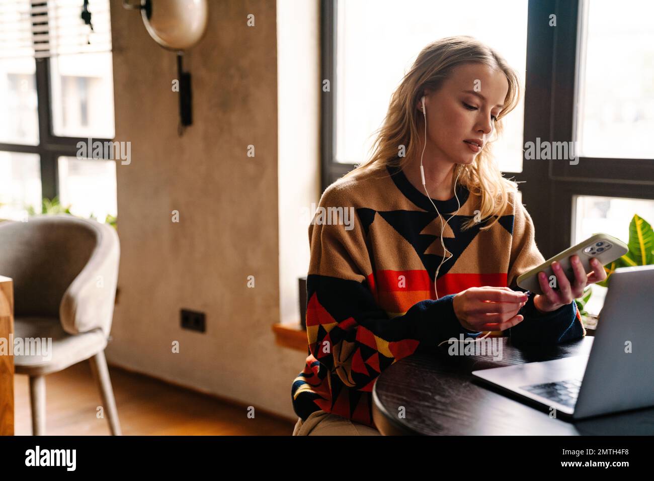 Giovane donna che indossa un pullover colorato che ricarica la batteria sul telefono cellulare mentre si siede al tavolo con un computer portatile Foto Stock