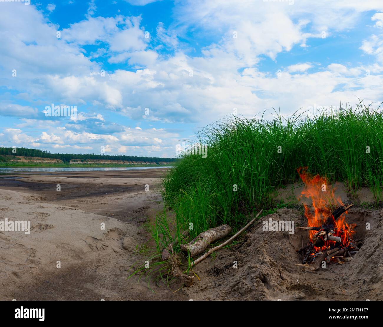 Un falò brucia durante il giorno vicino al fiume Vilyui nord a Yakutia nella sabbia sulla spiaggia accanto ad alta erba verde sotto un cielo blu e betulla Foto Stock
