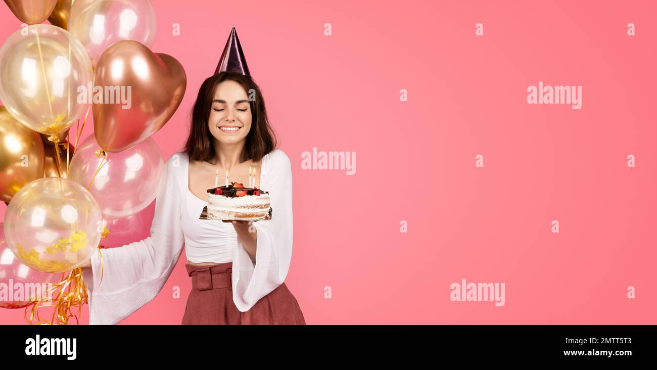 Allegra giovane donna caucasica in cappello tenere un sacco di palloncini gonfiabili, soffia le candele sulla torta Foto Stock