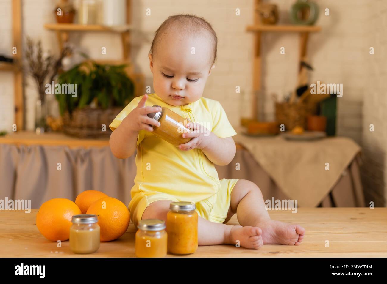 un capretto carino in un bodysuit giallo sta sedendosi nella cucina con le lattine di alimento del bambino. La prima alimentazione complementare del bambino. Puree per bambini. Alto qual Foto Stock