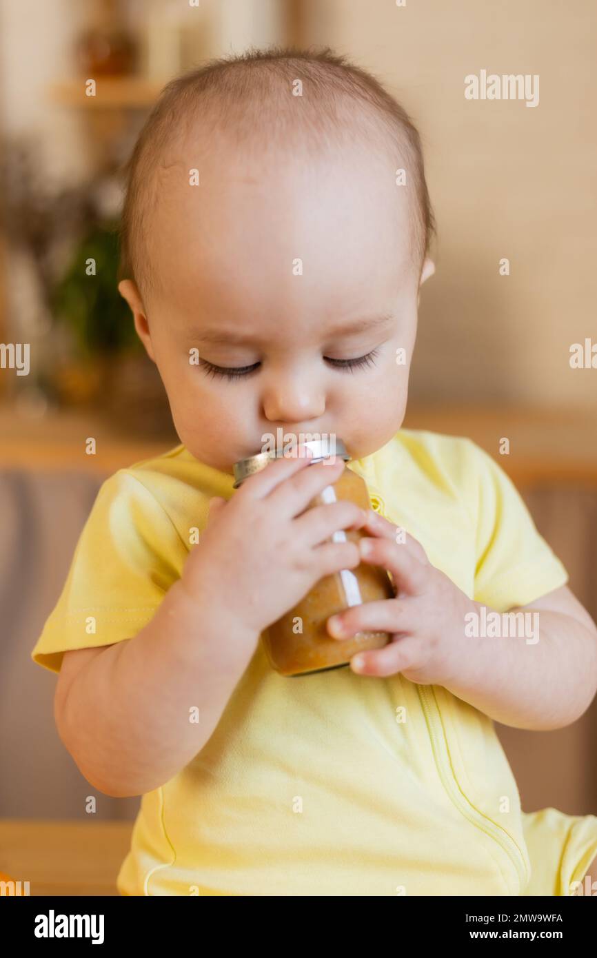 un capretto carino in un bodysuit giallo sta sedendosi nella cucina con le lattine di alimento del bambino. La prima alimentazione complementare del bambino. Puree per bambini. Alto qual Foto Stock