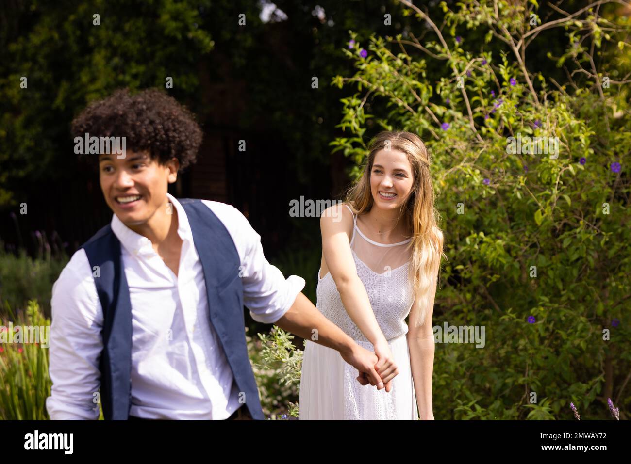 Felice sposa e sposo diverse tenendo le mani, camminando in giardino al matrimonio all'aperto Foto Stock