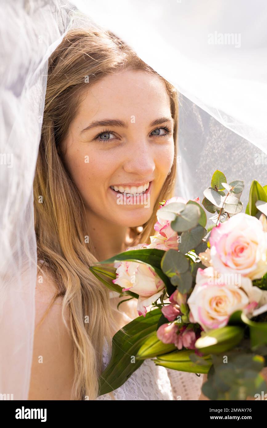 Ritratto verticale di sposa caucasica sorridente in velo che tiene bouquet all'aperto matrimonio Foto Stock
