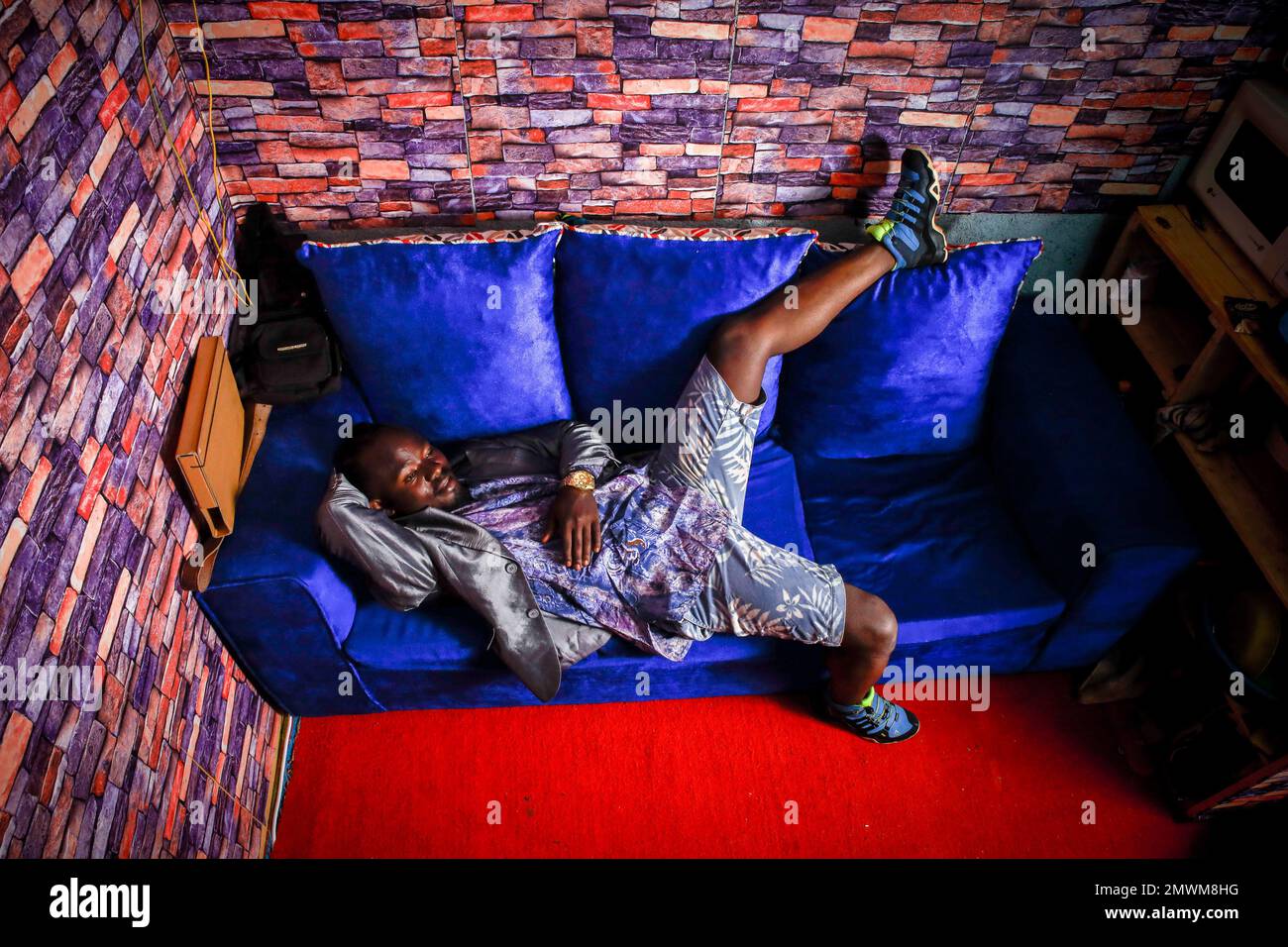 Clinton Chogo, di 23 anni, adagiato su una sedia all'interno della sua casa dal design colorato vestita con abiti di classe a Kibera. I giovani kenioti sono spinti in una competenza di avere le proprie case lontano dai genitori, dove soprattutto imparano ad affrontare la vita attraverso diverse sfide. La competizione di avere le camere meglio decorate piene di diversi abiti di design è un modo per tenere lontano dalle sfide quotidiane della vita, come l'abuso di droghe, le gravidanze adolescenziali matrimoni precoci, e giudizi negativi da parte della gente del posto. Per la maggior parte dei giovani avere una casa è l'inizio di una nuova vita Foto Stock