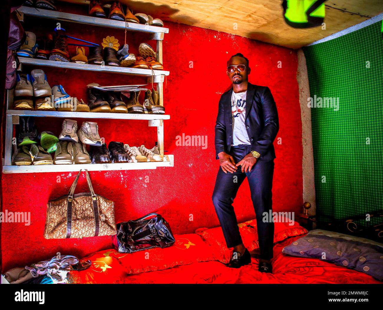Clinton Chogo, di 23 anni, si posa sul suo letto all'interno della sua casa dal design colorato vestita con abiti di classe a Kibera. I giovani kenioti sono spinti in una competenza di avere le proprie case lontano dai genitori, dove soprattutto imparano ad affrontare la vita attraverso diverse sfide. La competizione di avere le camere meglio decorate piene di diversi abiti di design è un modo per tenere lontano dalle sfide quotidiane della vita, come l'abuso di droghe, le gravidanze adolescenziali matrimoni precoci, e giudizi negativi da parte della gente del posto. Per la maggior parte dei giovani avere una casa è l'inizio di una nuova vita a. Foto Stock