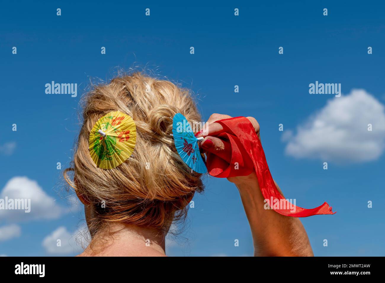 Una donna bionda dispone i capelli con due ombrelli di carta colorati e un nastro rosso, contro il cielo blu Foto Stock