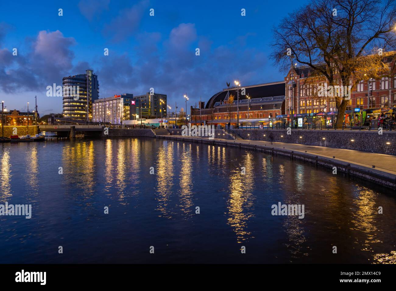 La parte anteriore della stazione centrale di Amsterdam IJzijde e i nuovi edifici a Stationsplein dall'altra parte del porto da Prins Hendrikkade Street al crepuscolo Foto Stock
