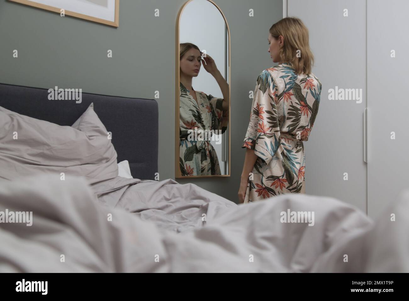 Giovane bella donna in accappatoio di seta in piedi nella parte anteriore dello specchio nella camera da letto Foto Stock
