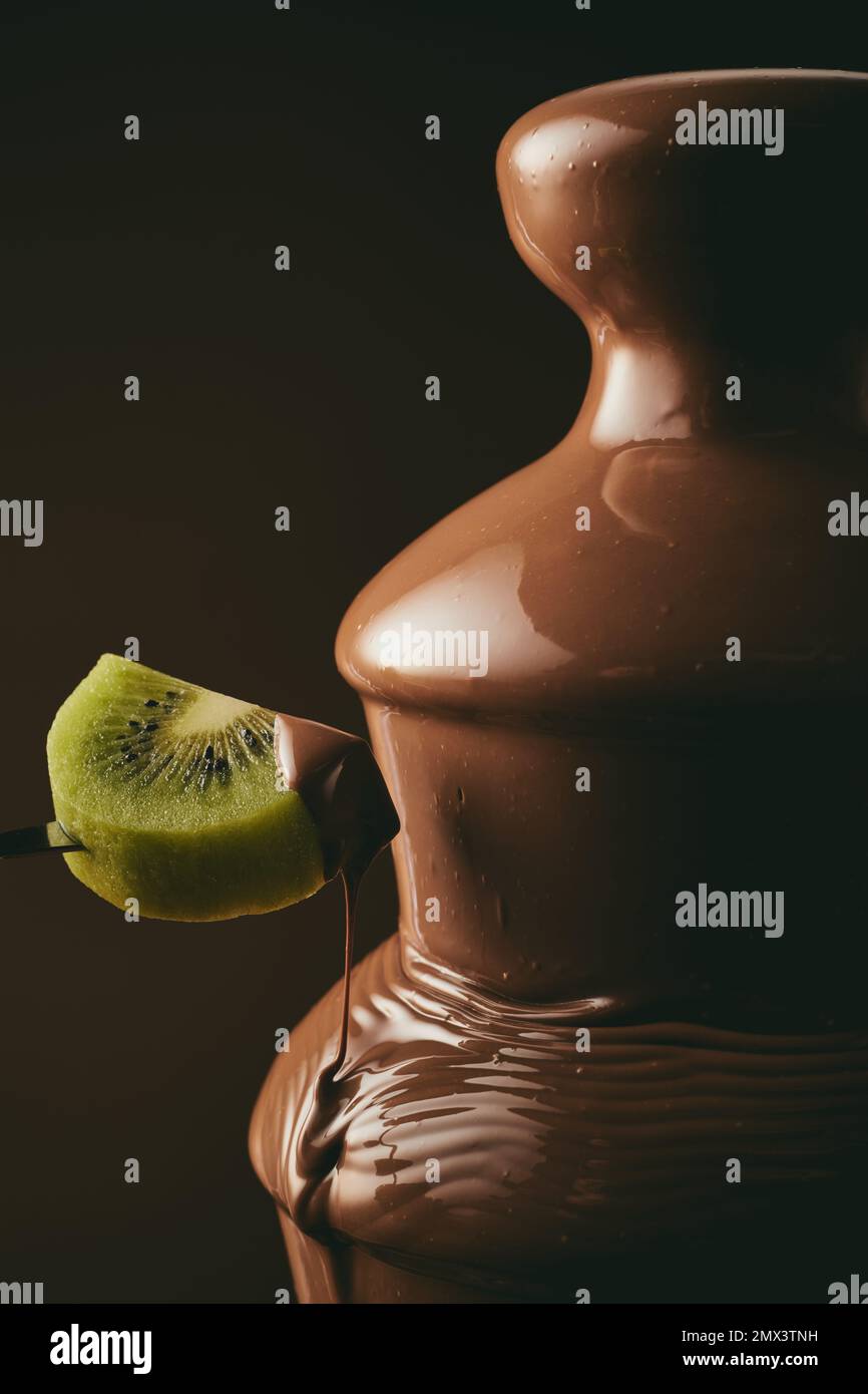 Persona irriconoscibile che immergerà un pezzo di kiwi dolce in una gustosa fontana di cioccolato durante la festa su sfondo nero Foto Stock
