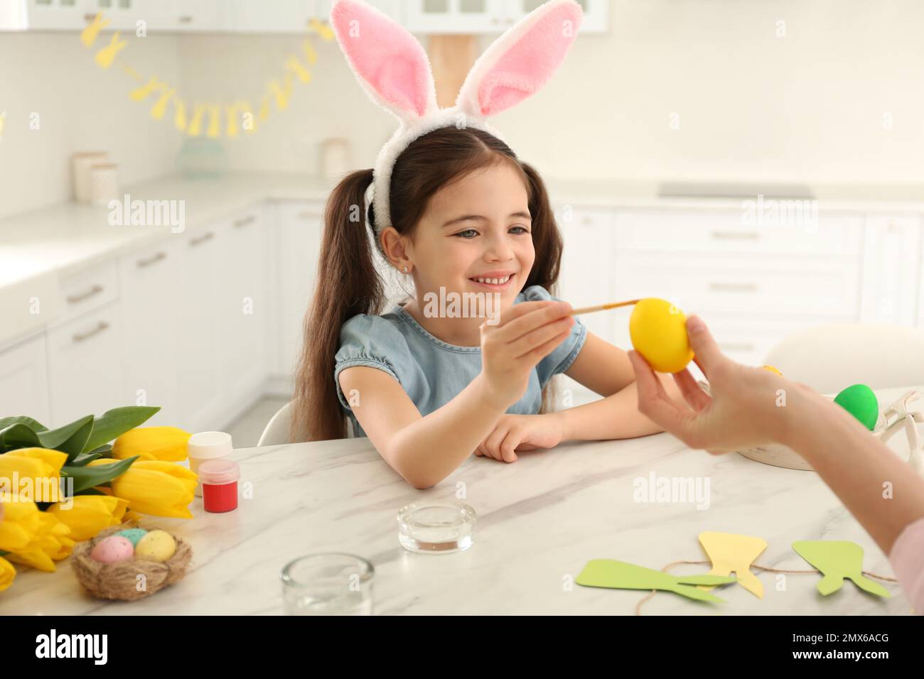 Ragazza piccola felice con coniglietto orecchie e sua madre pittura uovo di Pasqua in cucina Foto Stock