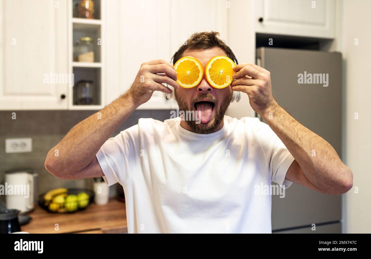 Uomo in piedi in cucina che copre gli occhi con fette di arancio e lingua attaccante fuori. Foto Stock