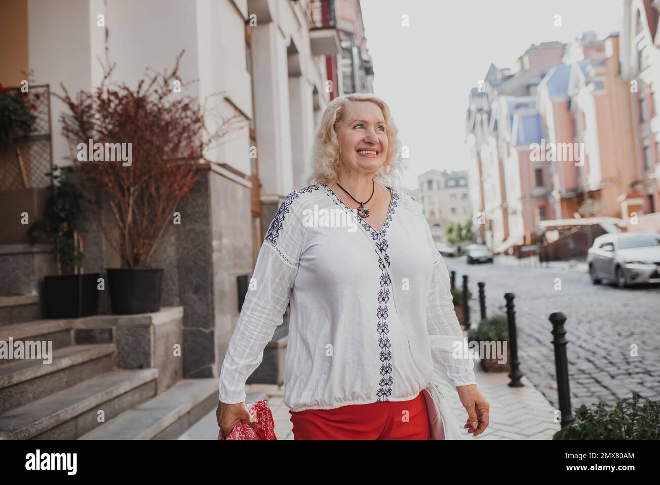 Ritratto di una donna matura felice che cammina su una strada cittadina in autunno. Bella donna con capelli biondi gode di una passeggiata durante il suo tempo libero in pensione Foto Stock