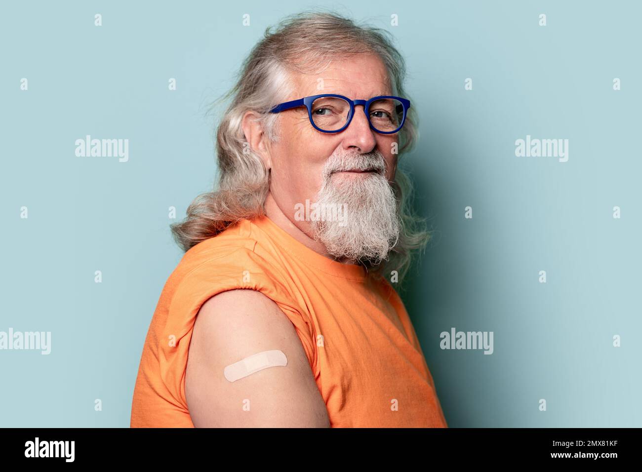 Uomo anziano in occhiali e t-shirt arancione con maniche arrotolate che mostrano bendaggio e guardare lontano su sfondo blu Foto Stock