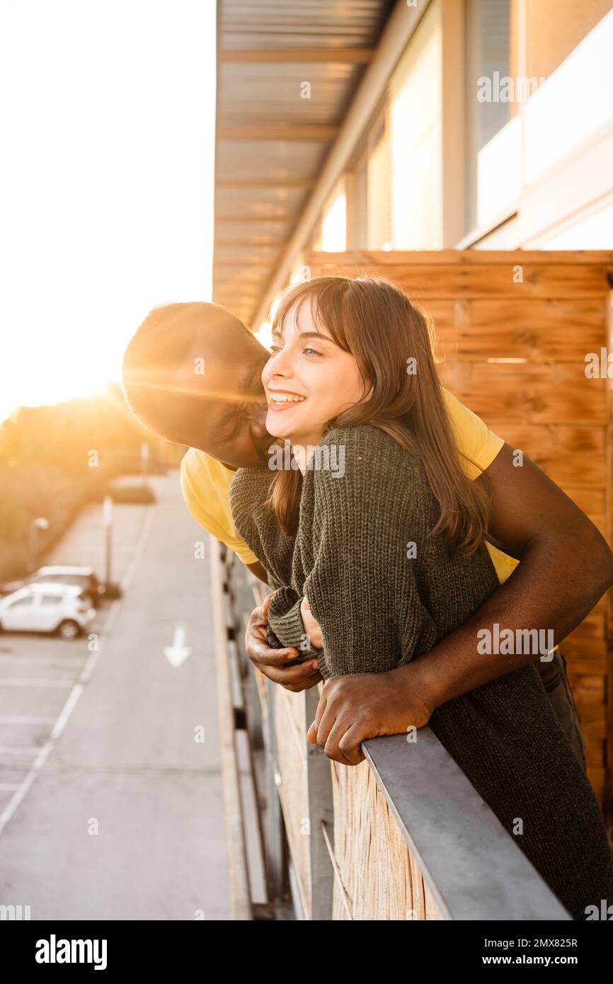 Vista laterale di una giovane coppia multirazziale deliziata in un abbigliamento casual sorridente e abbracciante mentre si alza in piedi sul balcone della casa e si appoggia sulla ringhiera contro s. Foto Stock