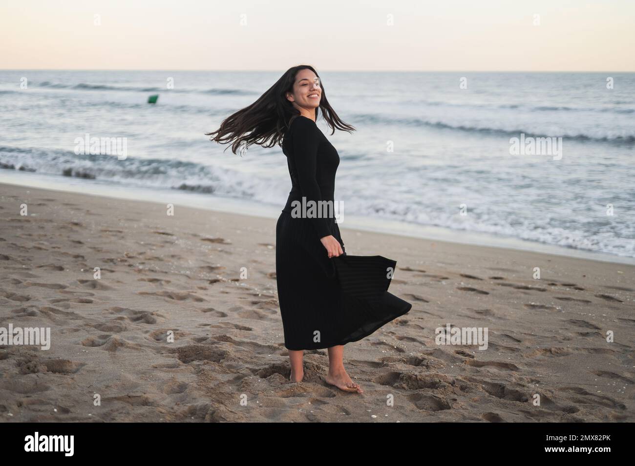 Vista laterale di sicuro giovane ragazza barefoted Ispanica turista in maxi nero vestito sorridente e scuotendo i capelli mentre si trova in piedi sulla spiaggia sabbiosa vicino a onde s. Foto Stock