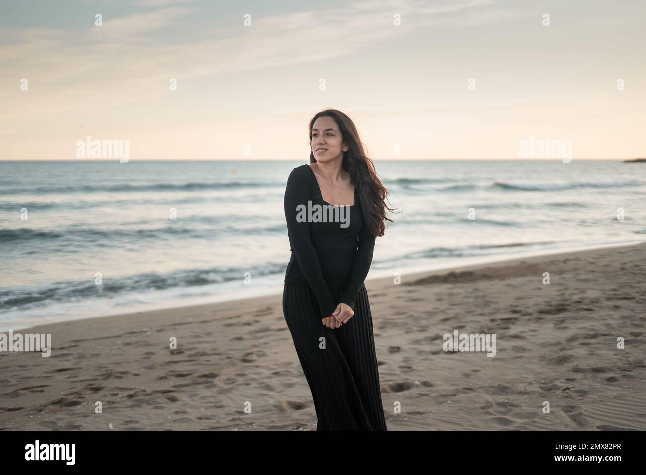Fiducioso giovane ragazza barefoted ispanica turista in maxi nero abito sorridente mentre si alza in piedi guardando lontano sulla spiaggia di sabbia vicino ondulato Foto Stock