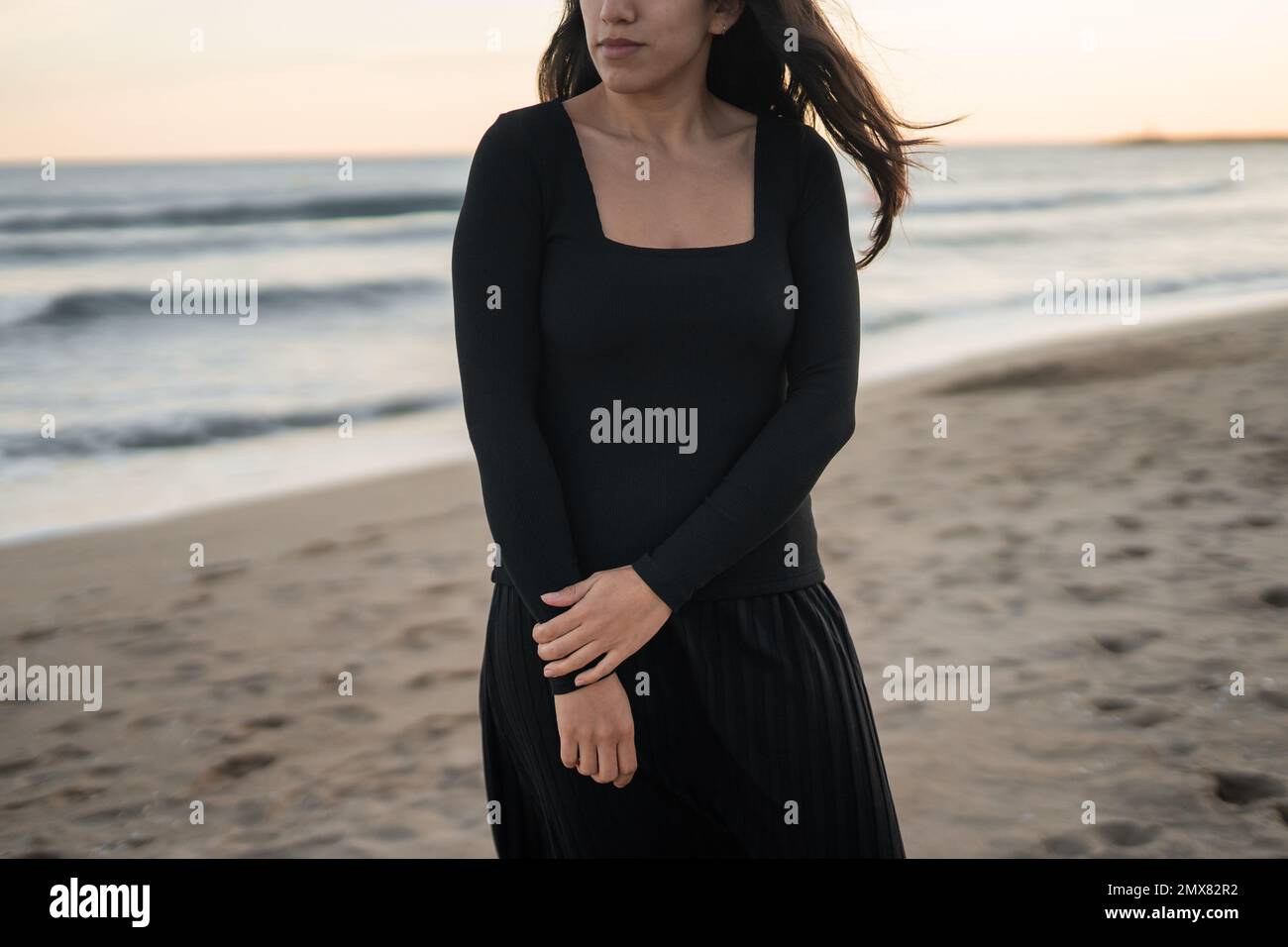 Corto irriconoscibile fiducioso giovane ragazza barefoted Ispanica turista in maxi vestito nero in piedi sulla spiaggia di sabbia vicino mare ondoso Foto Stock