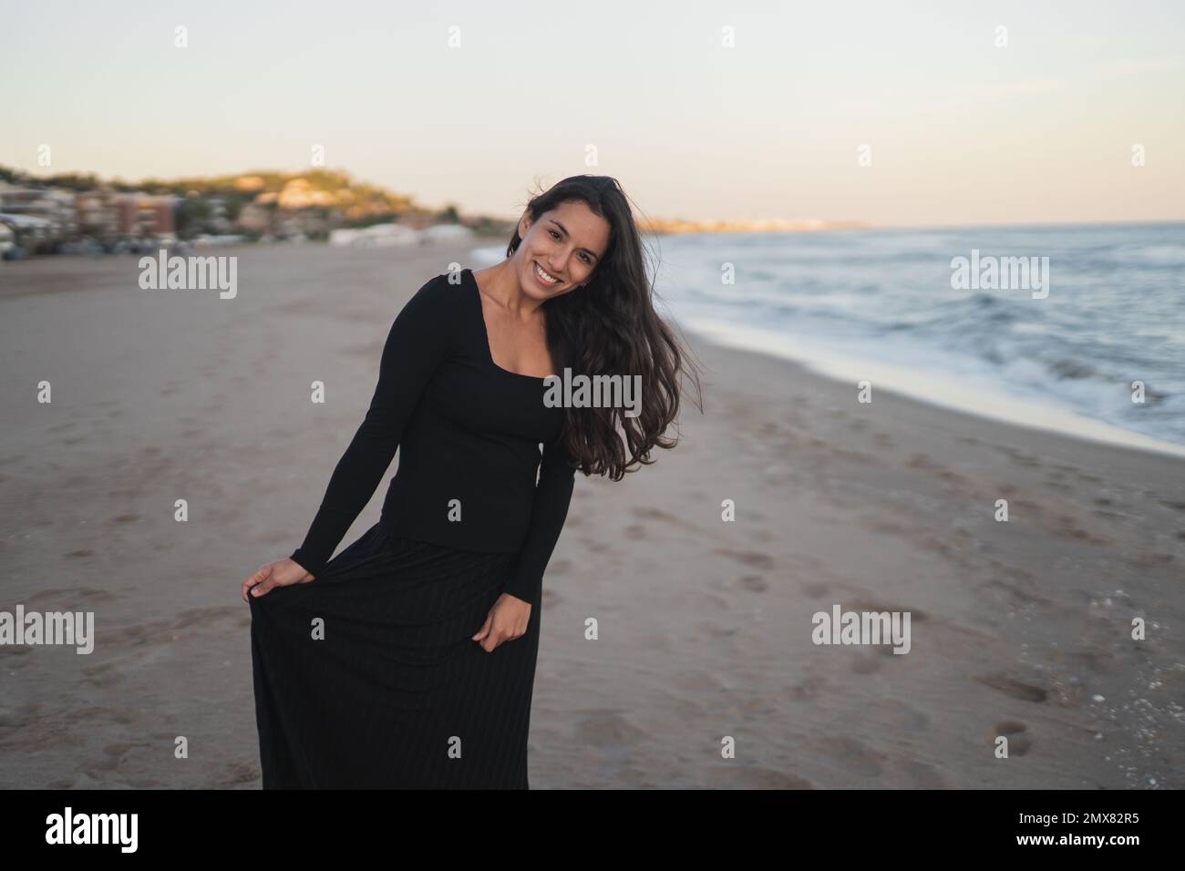 Fiducioso giovane ragazza barefoted ispanica turista in maxi nero abito sorridente mentre in piedi guardando la macchina fotografica sulla spiaggia di sabbia vicino mare ondulato sotto il sole Foto Stock