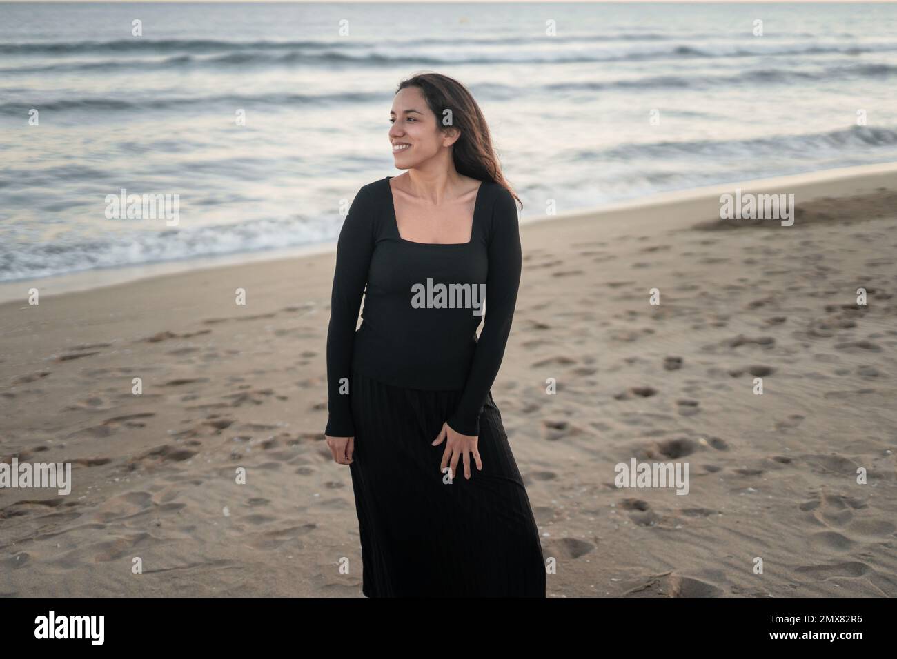 Fiducioso giovane ragazza barefoted ispanica turista in maxi nero abito sorridente mentre si alza in piedi guardando lontano sulla spiaggia di sabbia vicino ondulato Foto Stock