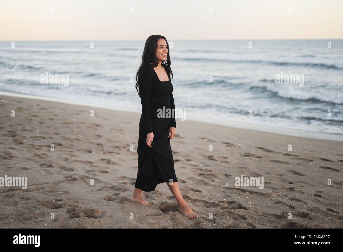 Vista laterale di sicuro giovane ragazza barefoted Ispanica turista in maxi nero vestito sorridente e scuotendo i capelli mentre si trova in piedi sulla spiaggia sabbiosa vicino a onde s. Foto Stock