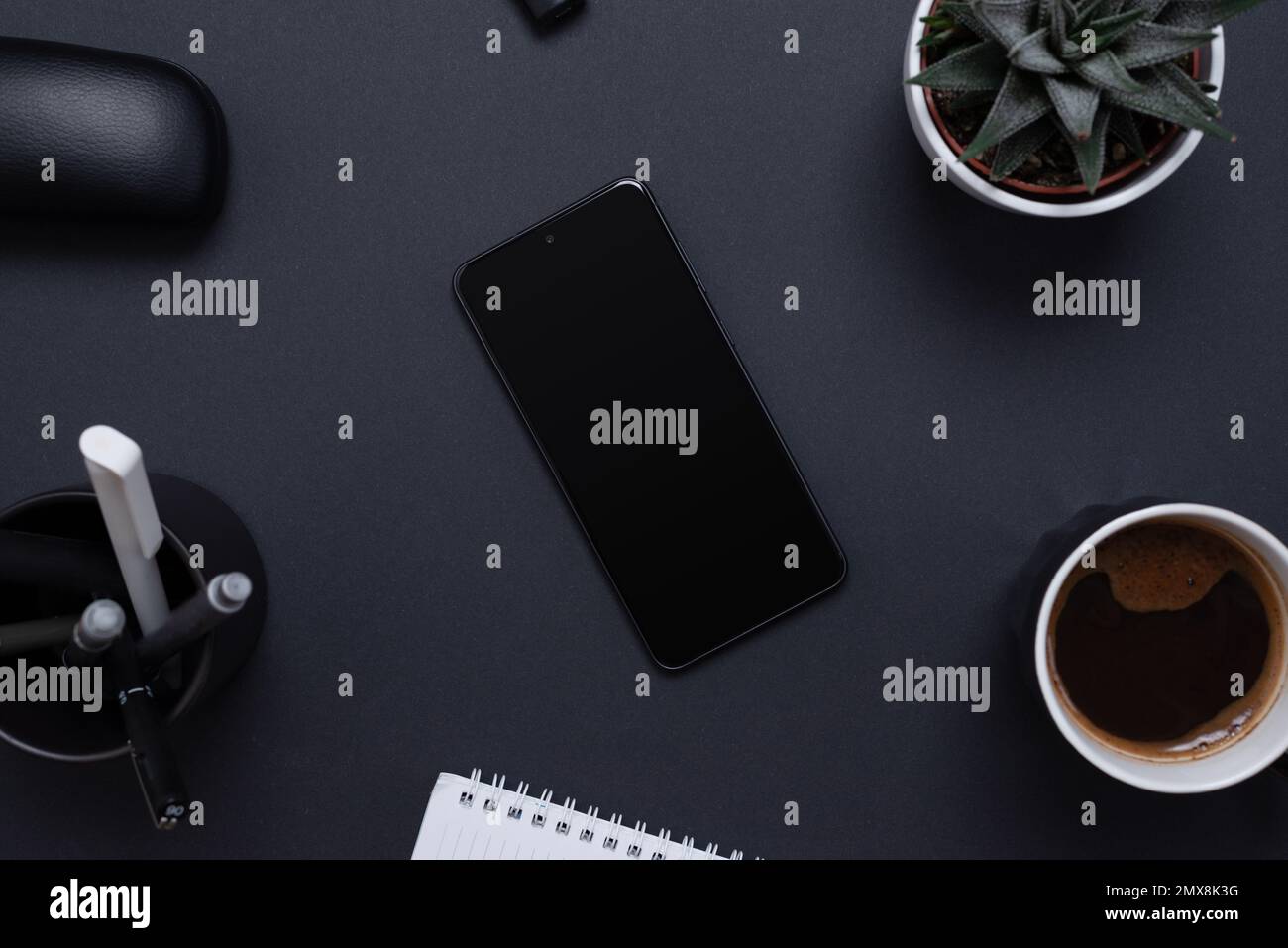 Scrivania minimalista e organizzata con uno smartphone vuoto circondato da una pianta, penne, caffè e blocco note. Schermata vuota per la presentazione dell'app mockup. Foto Stock