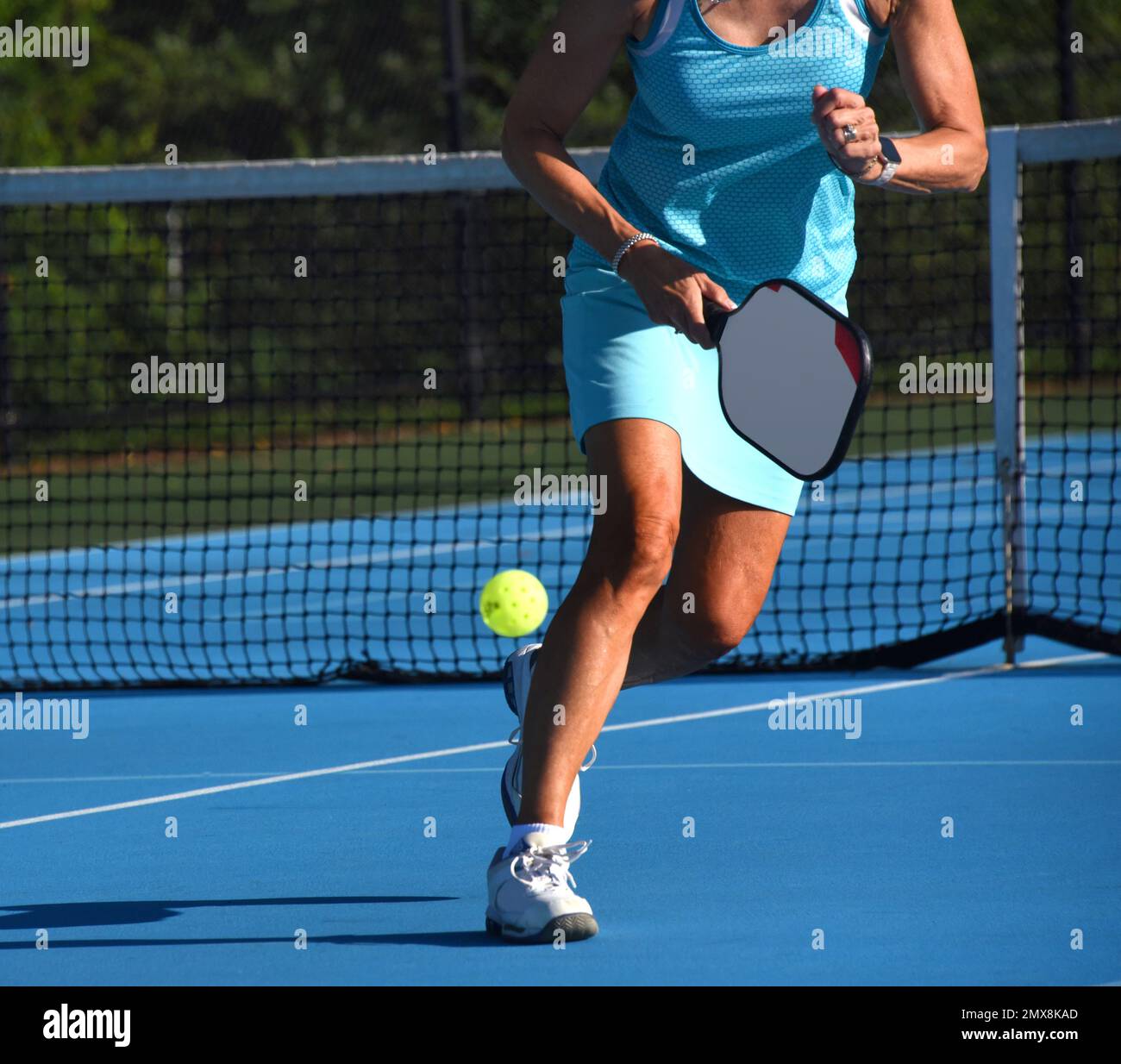Il giocatore di palla di pickle senior corre per intercettare una palla di pickle gialla. È femminile e indossa un colorato vestito da tennis turchese. Foto Stock