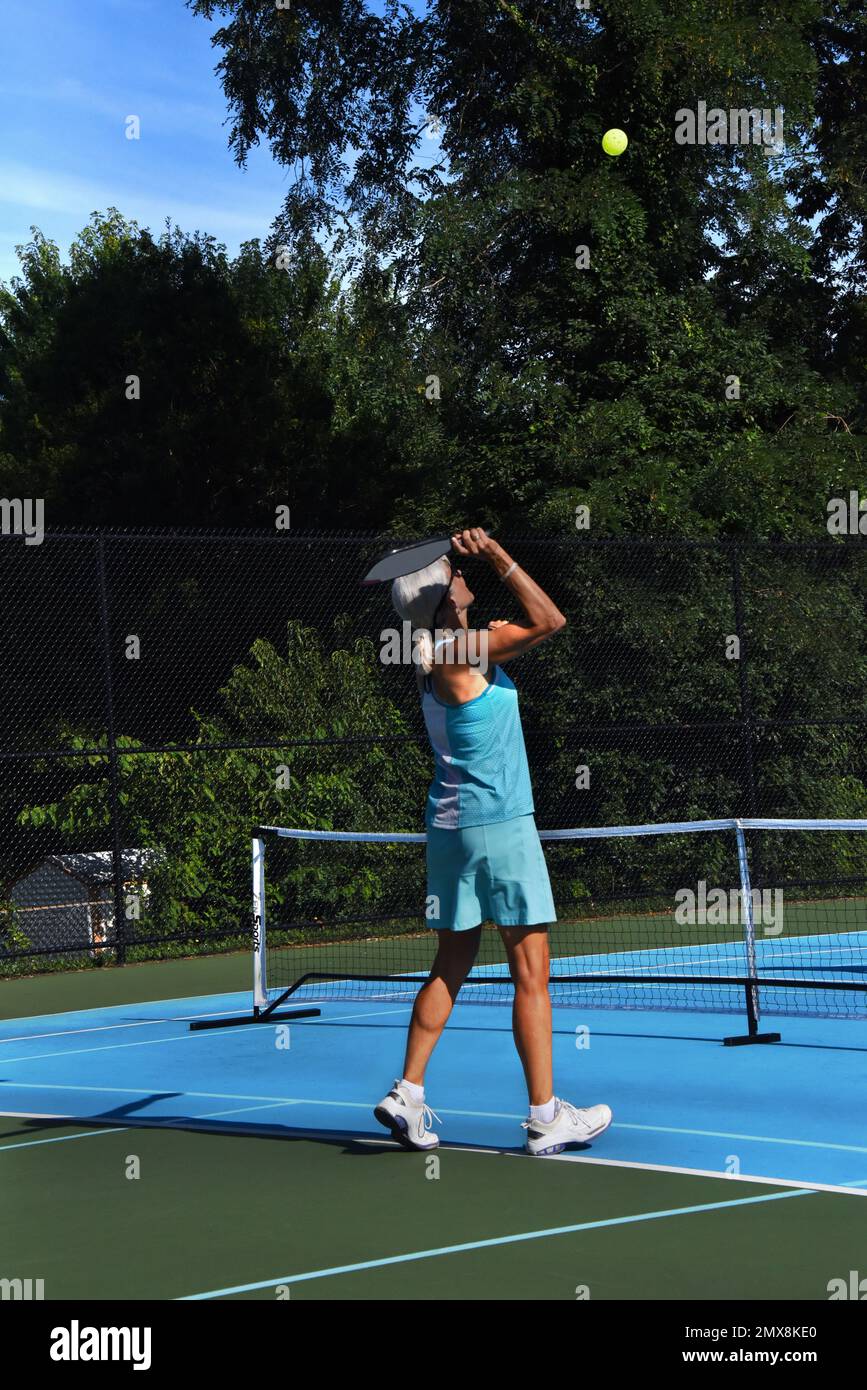 Il giocatore senior, femminile, di pickle ball si prepara a colpire un pickle ball alto. La sua concentrazione è concentrata sul suo swing e ritorno. Indossa un turquo Foto Stock