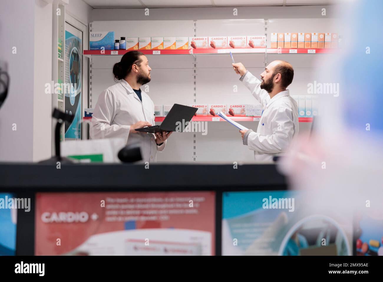 Due farmacisti uomini che fanno l'inventario medicamenti che controllano i pacchetti di farmaci, digitando le informazioni sulle vitamine sul portatile. La farmacia è una posizione affidabile e conveniente per riempire il farmaco di prescrizione Foto Stock