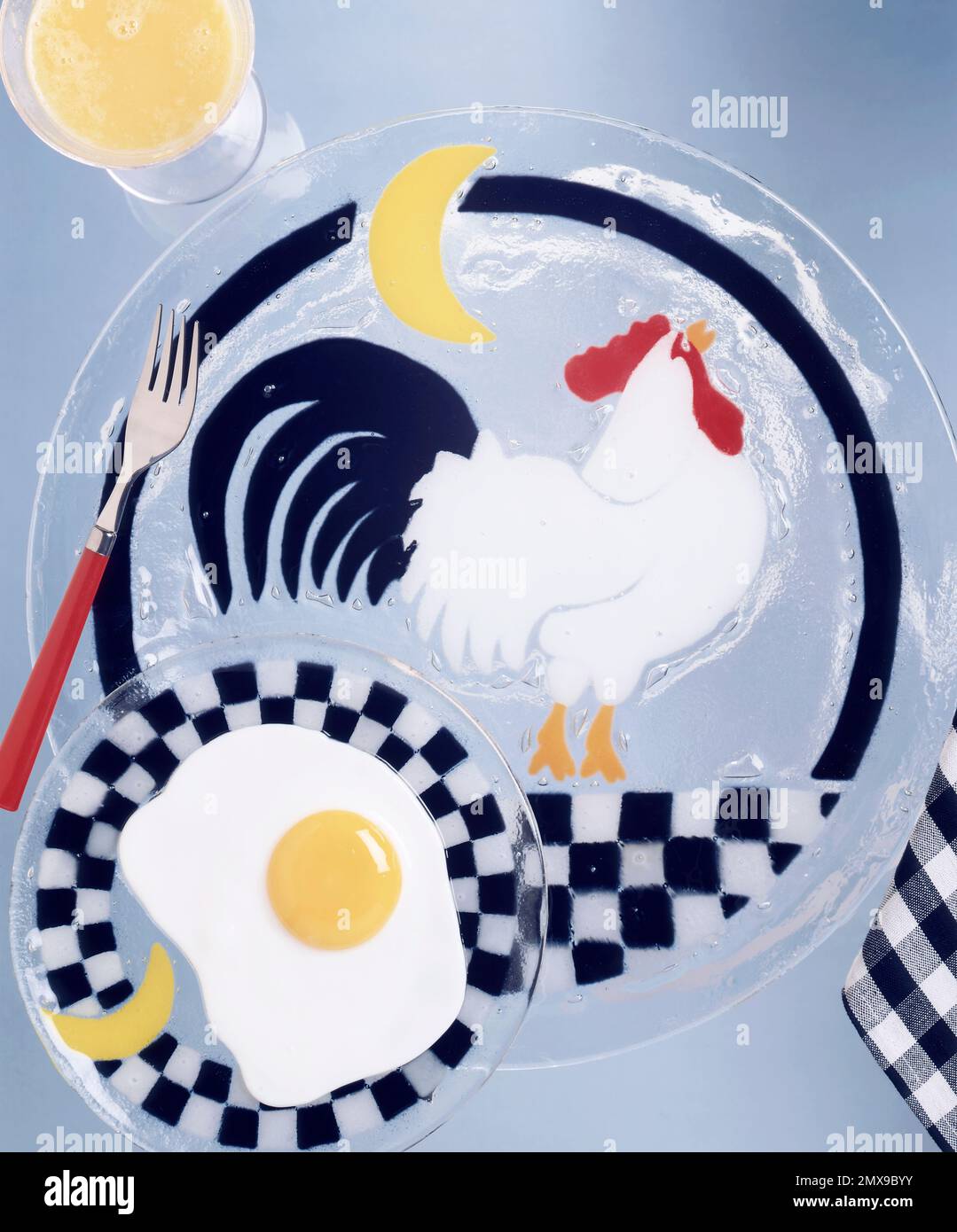 Composizione bizzarra di un uovo fritto con un piatto di gallo di vetro. Fotografia verticale a colori in studio. Immagine concettuale per la colazione, con bicchiere di succo di frutta Foto Stock