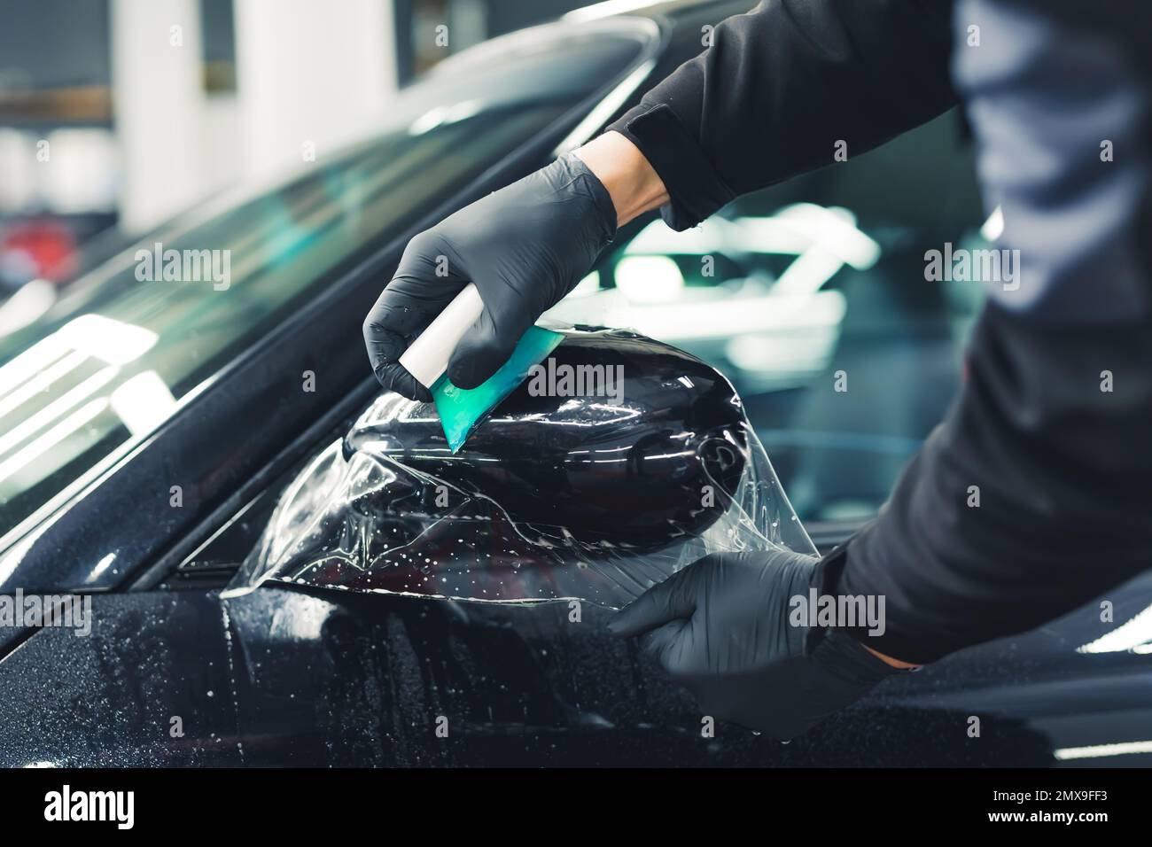 Primo piano interno dell'installazione della pellicola di protezione della vernice sullo specchio laterale di un'auto di lusso moderna. Persona non riconoscibile che detiene attrezzature specialistiche. Foto di alta qualità Foto Stock