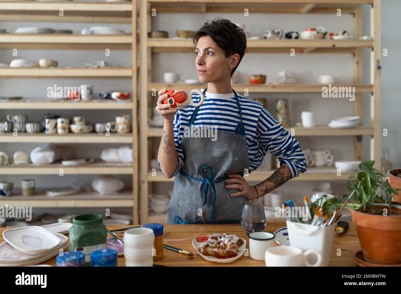 Donna ceramista creativa in grembiule sporco godendo il suo caffè mattutino dalla tazza di argilla e guardando via Foto Stock