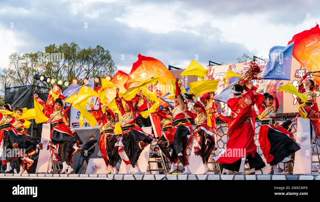 La squadra giapponese di ballerini Yosakoi ballare e cantare mentre sventola bandiere gialle sul palco all'aperto al festival Kyusyu Gassai di Kumamoto. Crepuscolo. Foto Stock