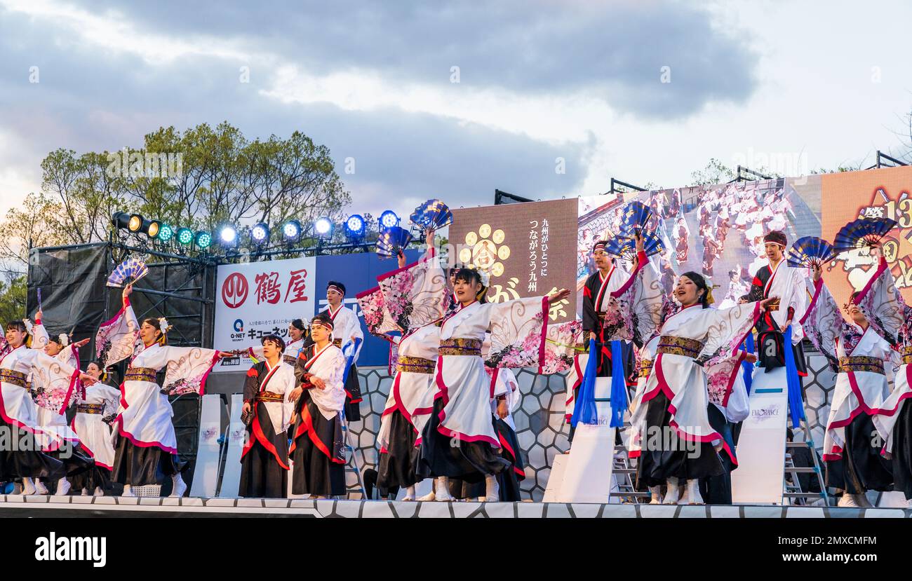 Squadra giapponese di danzatrici Yosakoi che ballano e tengono naruko e tifosi sul palco all'aperto al festival Kyusyu Gassai di Kumamoto. Crepuscolo. Foto Stock