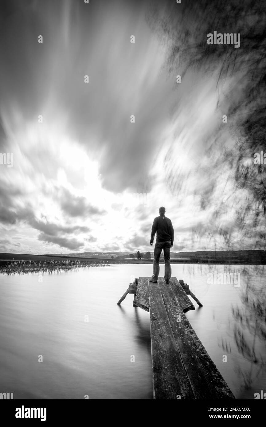 Un'immagine verticale in scala di grigi di una persona su una passerella vicino al lago Foto Stock