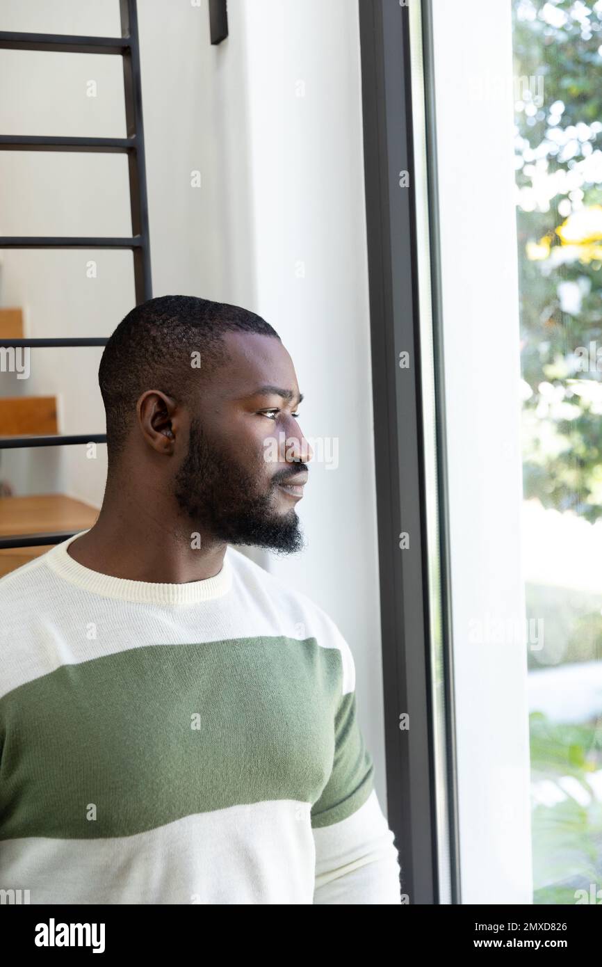 Verticale di un uomo afro-americano ponderato in piedi, guardando fuori dalla finestra del soggiorno, copia spazio Foto Stock