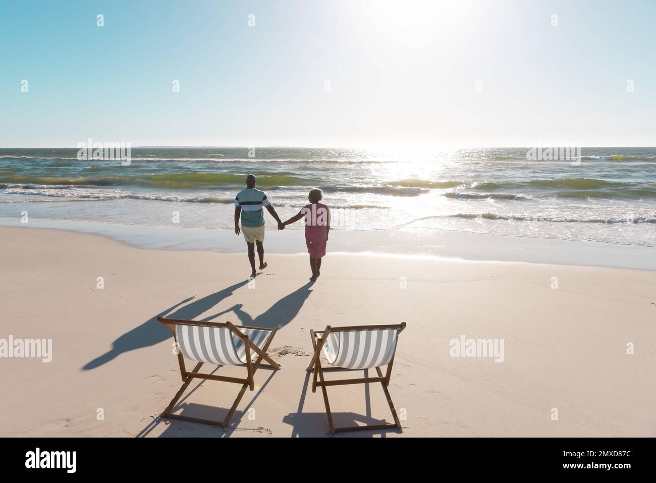 Vista posteriore della coppia anziana afro-americana che tiene le mani e cammina verso il mare sotto il cielo Foto Stock