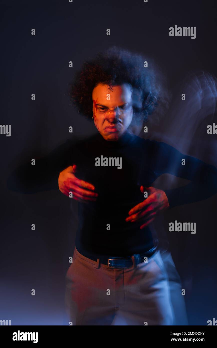 sfocatura del movimento di aggressivo afro-americano con disturbo bipolare e volto sanguinoso guardando la fotocamera al buio con luce arancione e blu, immagine stock Foto Stock