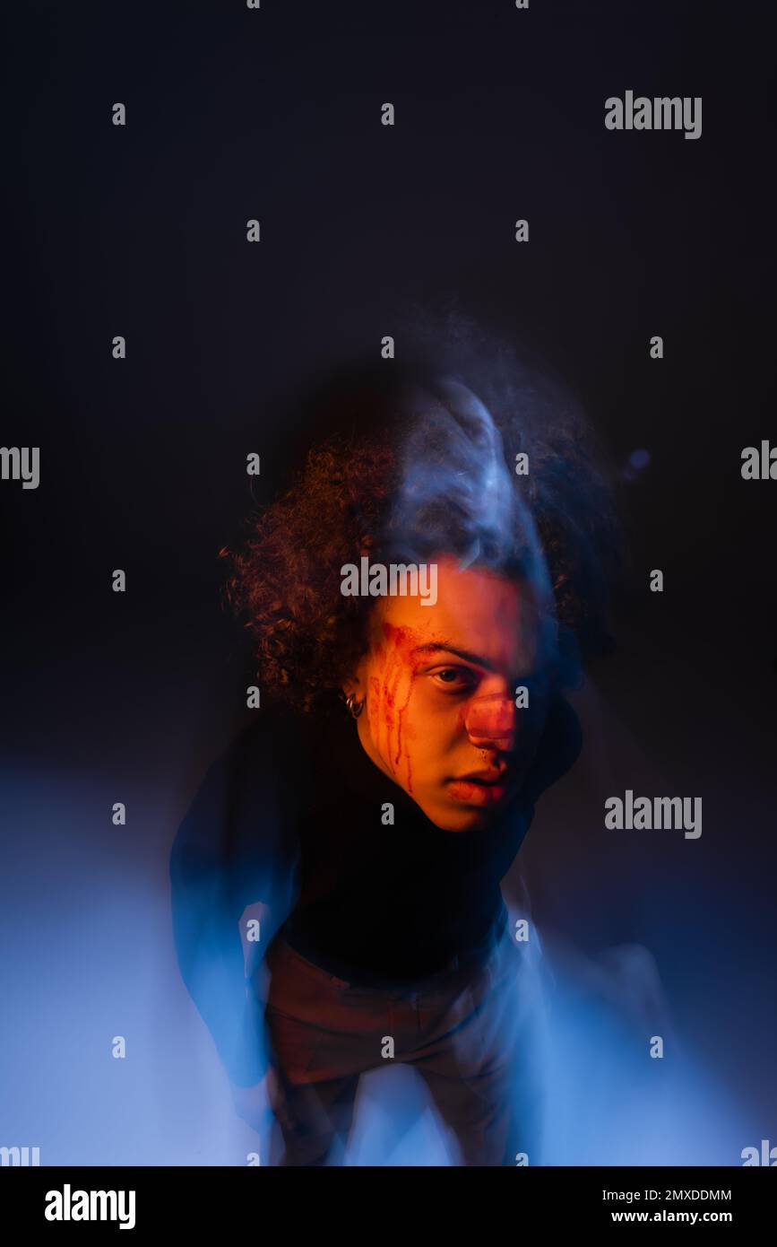doppia esposizione dell'uomo afroamericano con disturbo bipolare e faccia sanguinante guardando la fotocamera in nero con luce arancione e blu, immagine stock Foto Stock
