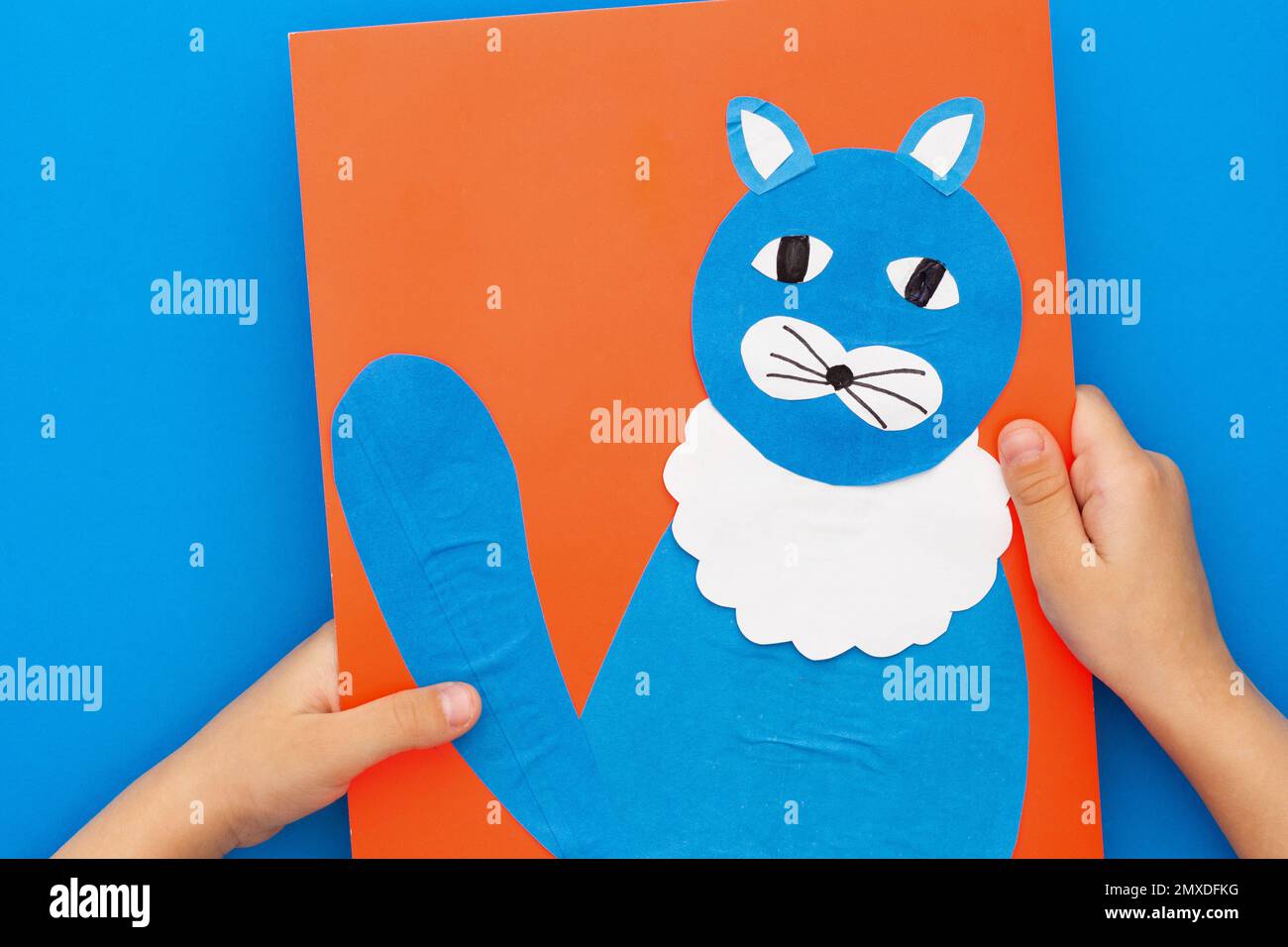 Vista dall'alto delle mani dei bambini che tengono gatto di carta artigianale su foglio rosso su sfondo blu Foto Stock