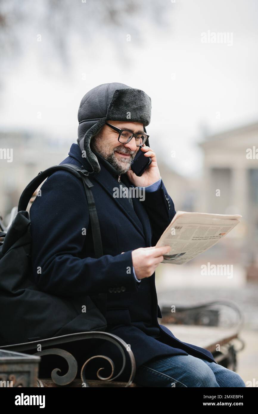 Un uomo anziano gode di una fredda mattinata invernale in città. Un uomo siede su una panchina nella piazza, legge un giornale e chiacchiera allegro con gli amici su un Foto Stock