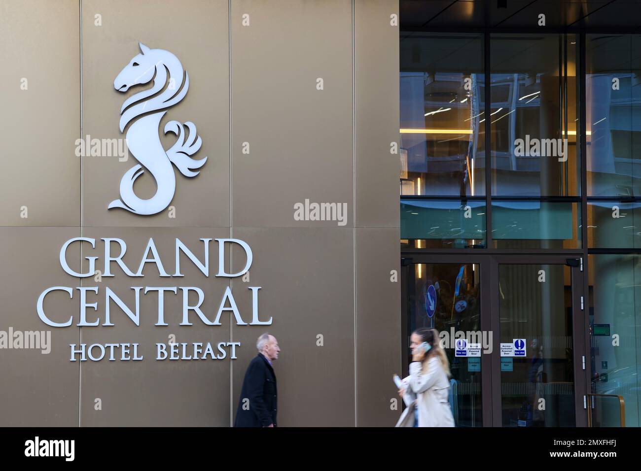 Vista sulla strada dell'ingresso principale del Grand Central Hotel Belfast con due pedoni che passano davanti al cartello del Grand Central Hotel Belfast. Foto Stock