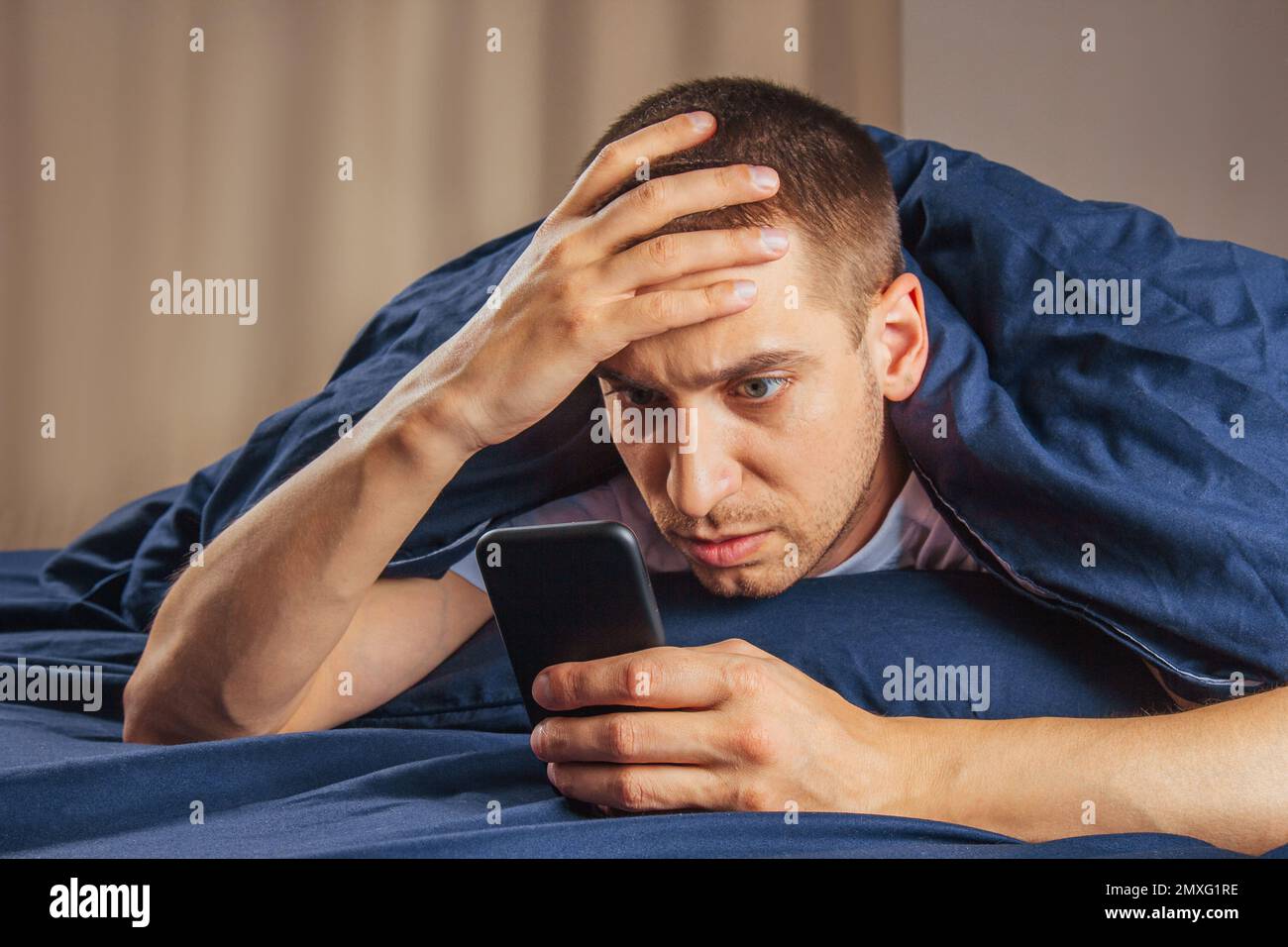 Cattive notizie, problemi, offerte e pubblicità. Scioccato ragazzo attraente sconvolto legge un messaggio sul telefono e preme la mano al suo volto su un letto bianco Foto Stock