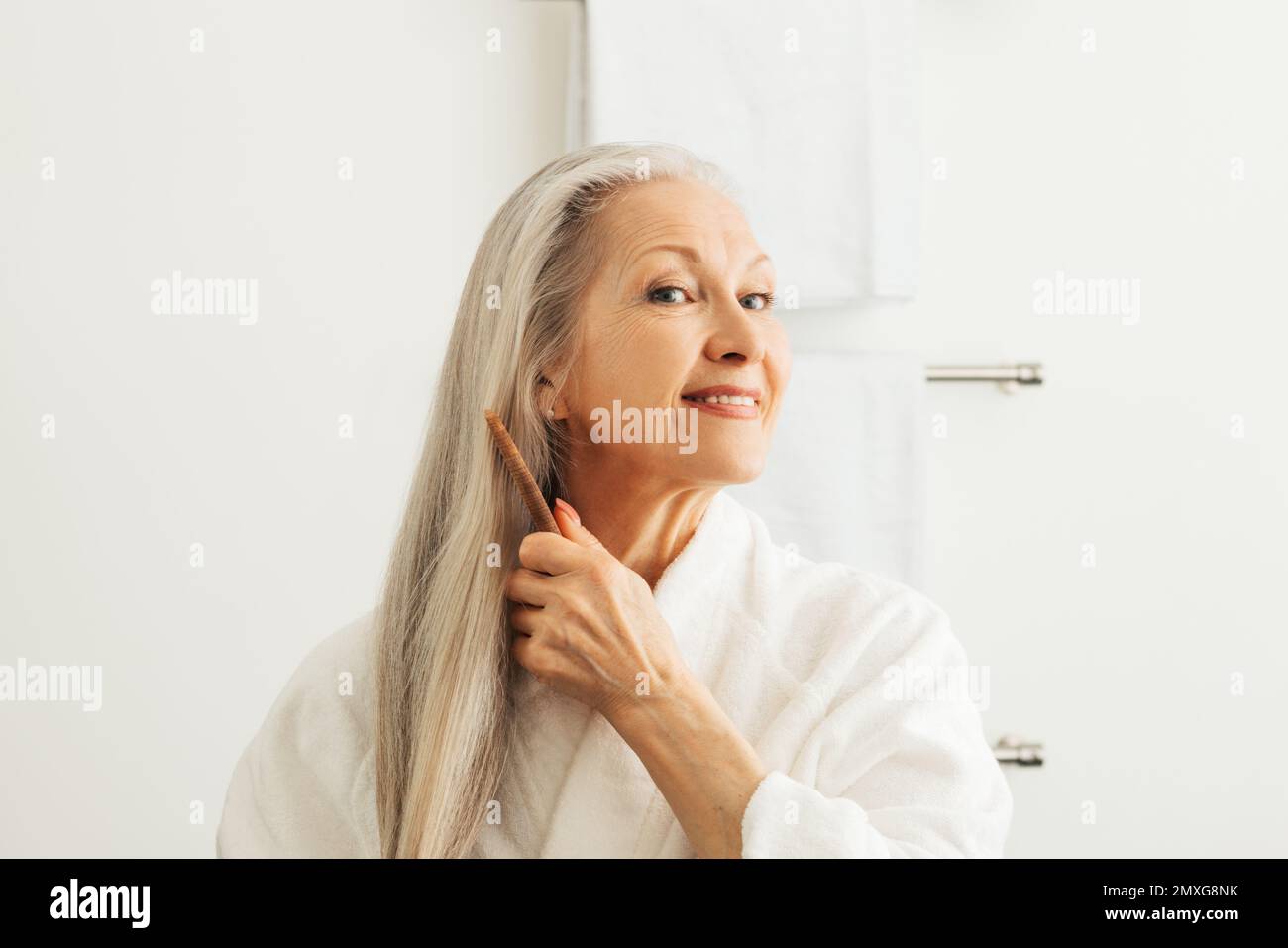 Donna adulta anziana che pettina i capelli con un pettine di legno. Donna sorridente in un accappatoio che si prende cura dei suoi lunghi capelli grigi. Foto Stock
